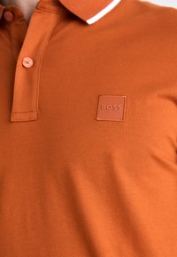 Trumpas rankovėmis polo marškinėliai deginto oranžinio audinio, turintys klasikinį apykaklę, dvi mygtukus ir kvadratinę gumos logotipo etikete "BOSS."