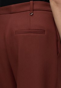 Gros plan sur un pantalon marron texturé avec une poche passepoilée au dos et une passants de ceinture sur la hanche et le côté d’une personne.