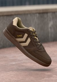 Sneaker in suede marrone con dettagli beige e una finitura testurizzata. Presenta una punta arrotondata, lacci piatti e un logo sul lato.