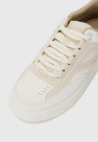 Sneaker bianco e beige chiaro con tomaia in pelle testurizzata, punta tonda e suola spessa in gomma. Presenta lacci in cotone piatti e dettagli perforati.