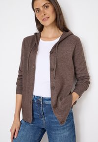Brauner Strickcardigan mit Kapuze, Knopfverschluss und gerippten Bündchen. Getragen über einem weißen Shirt und kombiniert mit blauen Jeans.