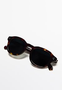 Zonnebril met schildpadmontuur en zwarte glazen, rond van vorm, opvallende metalen accenten naast de glazen en dikke pootjes voor stevigheid.