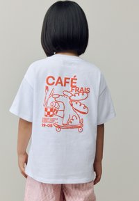 T-shirt en coton blanc avec une illustration graphique orange vif d'un chien sur un skateboard accompagné de café et de pâtisseries. Le texte inclut "CAFÉ FRAIS."