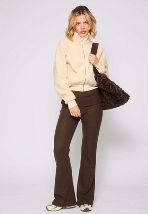 Vrouw in een beige ritssluiting fleecejack en bruine flared broek met een grote donkerbruin gestructureerde schoudertas, staande tegen een effen achtergrond.