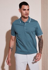 Turkizna polo majica z belim ovratnikom in poudarki na rokavih, kratkimi rokavi in gumbom. Nosena z svetlimi hlačami in prikazuje tetovaže.