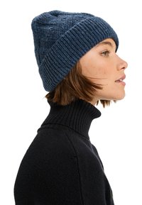 Strikket hue i mørkeblå med et struktureret mønster og ribbet kant. Båret med en sort turtleneck sweater, set fra siden.