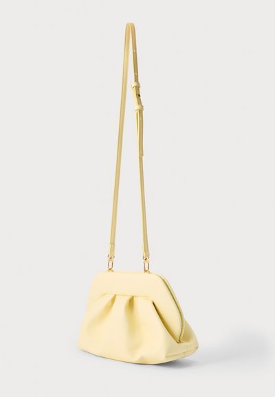 Petit sac en cuir jaune doux avec un détail froncé et une longue bandoulière réglable suspendu sur un fond blanc.