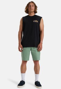 Black sleeveless t-shirt with graphic print on the chest, paired with green shorts. Model wears white socks and navy sneakers.