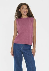 Frau mit lockigem Haar, die ein ärmelloses pinkes Oberteil und blue Jeans trägt, lächelt und steht vor einem weißen Hintergrund.