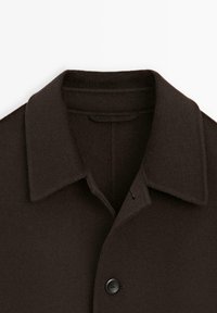 Cappotto di lana marrone con colletto classico, chiusura con un singolo bottone nero e texture morbida. Dettagli accentuati sul collo e sulla parte anteriore superiore.