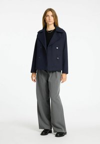 Cappotto navy doppiopetto con ampi risvolti, realizzato in un tessuto liscio, abbinato a pantaloni grigi a pieghe e scarpe nere.
