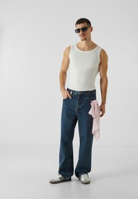 Hvit ribbet tank topp, oversized blå denim jeans, grå og hvite sneakers, og et rosa bandana knyttet i livet; solbriller brukt.