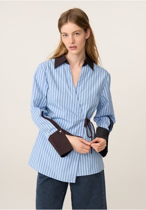 Jonge vrouw die een blauw gestreepte overslagblouse met contrasterende donkerbruine kraag en manchetten verstelt, draagt een donkere broek tegen een effen achtergrond.