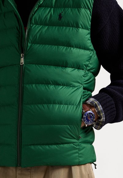Green puffer vest with quilted design and full zipper. A navy sweater is worn underneath, and a blue watch is visible on the wrist.
