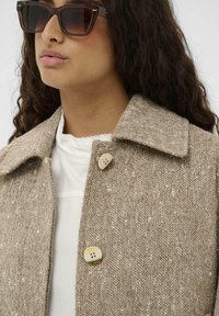 Bež jakna s ribjo vzorcem in ovratnikom ter z enim gumbom, izdelana iz teksturirane volnene tkanine. Model nosi sončna očala.