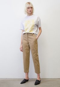 Bílý grafický tričko s žlutým designem, béžové kalhoty s vysokým pasem a černé slip-on boty. Hladká látka s uvolněným střihem.