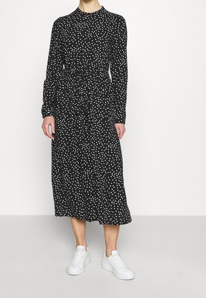 Kobieta ubrana w czarną sukienkę midi z długim rękawem w białe kropki i białe trampki, stojąca na jasnym, jednolitym tle.