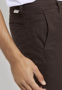 Pantaloni in cotone marrone scuro con taglio slim, caratterizzati da una tasca laterale e dettagli cuciti. Etichetta visibile sulla vita. Mano appoggiata vicino ai pantaloni.