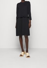 Svart klänning med långa ärmar och volangdetalj vid midjan, tillsammans med nudefärgade öppna klackar med tjock sula. Slätt tyg, knälång design.