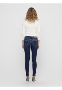 Úzké džíny v tmavě modrém denimu s vysokým pasem a nášivkou na zadní straně. Spárováno s přiléhavým bílým topem s dlouhým rukávem a béžovými podpatky.