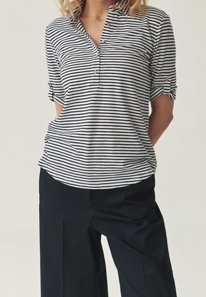 Nainen, joka pukeutunut mustavalkoraidalliseen, kolmen neljäsosan hihaiseen puseroon, jossa on napit, ja mustiin leveälahkeisiin housuihin, kädet selän takana.