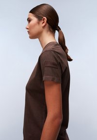 T-shirt in cotone marrone con scollo tondo, maniche corte e un patch logo in rilievo sulla spalla sinistra. Texture liscia.