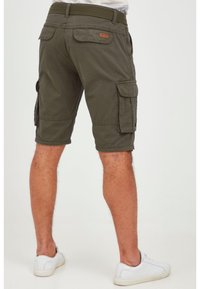 Man wearing olive green cargo shorts with multiple pockets and white sneakers, standing against a plain light background.