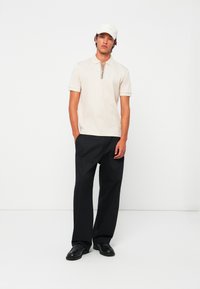 Ung mann iført en beige polo skjorte, svarte vide bukser, svarte sko og en beige caps, stående mot en hvit bakgrunn.