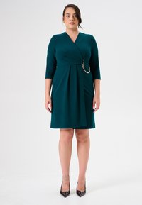 Tee-farbenes Wickelkleid mit drei Viertelärmeln, das eine drapierte Vorderseite, ein kreisförmiges Metallelement und einen knielangen Saum aufweist. Glatter Stoff.