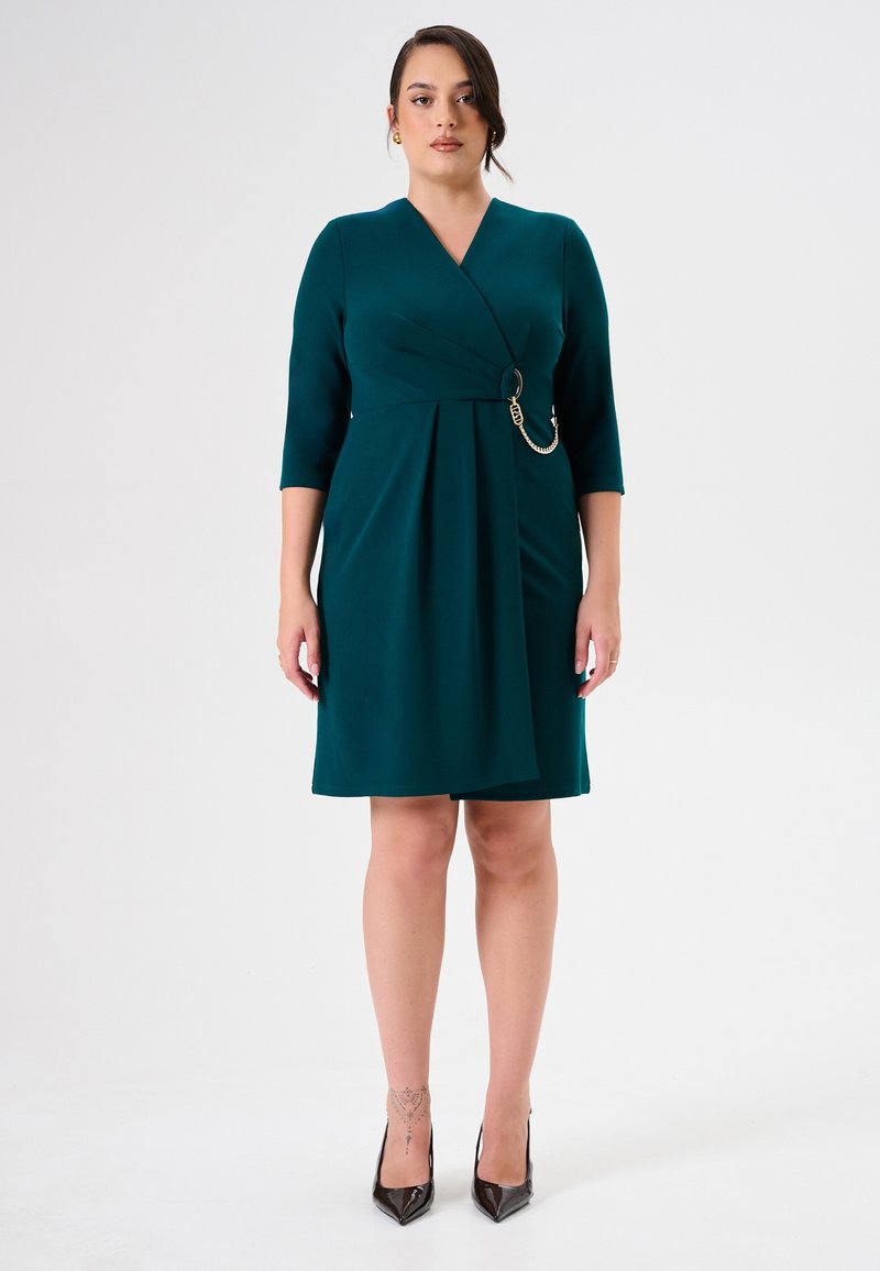Tee-farbenes Wickelkleid mit drei Viertelärmeln, das eine drapierte Vorderseite, ein kreisförmiges Metallelement und einen knielangen Saum aufweist. Glatter Stoff.