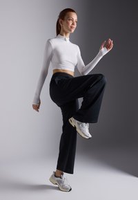 Bijela majica dugih rukava za sport, crne široke hlače i sivi New Balance tenisice. Model pozira s jednom nogom podignutom i ispruženom rukom.