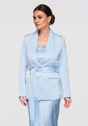 Donna con chignon liscio che indossa un blazer azzurro chiaro con cintura sopra un vestito testurizzato e orecchini circolari, rivolta verso destra su uno sfondo semplice.