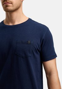 Marineblaues T-Shirt mit kurzen Ärmeln, aus weicher Baumwolle, mit einer Brusttasche auf der linken Seite und einem Rundhalsausschnitt mit minimalen Nähdetails.