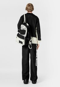 Mochila en blanco y negro con múltiples bolsillos, correas ajustables y un exterior con textura. El modelo lleva pantalones negros y una camiseta negra.