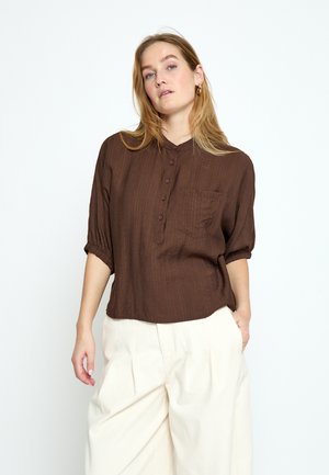 Kobieta ubrana w luźną brązową bluzkę na guziki z kieszenią oraz luźne białe plisowane spodnie, stojąca na jednolitym jasnym tle.