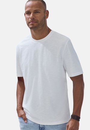 Mann mit kurzen Haaren und Bart, der ein schlichtes weißes kurzärmeliges T-Shirt und hellblaue Jeans trägt, steht mit einer Hand in der Tasche.