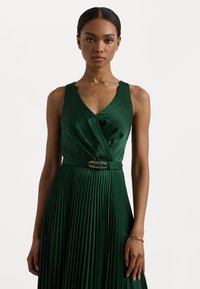 Grön plisserad klänning med djup V-ringning, satinmaterial och ett dekorativt bälte med en silverknäppning. Slät textur och elegant silhuett.