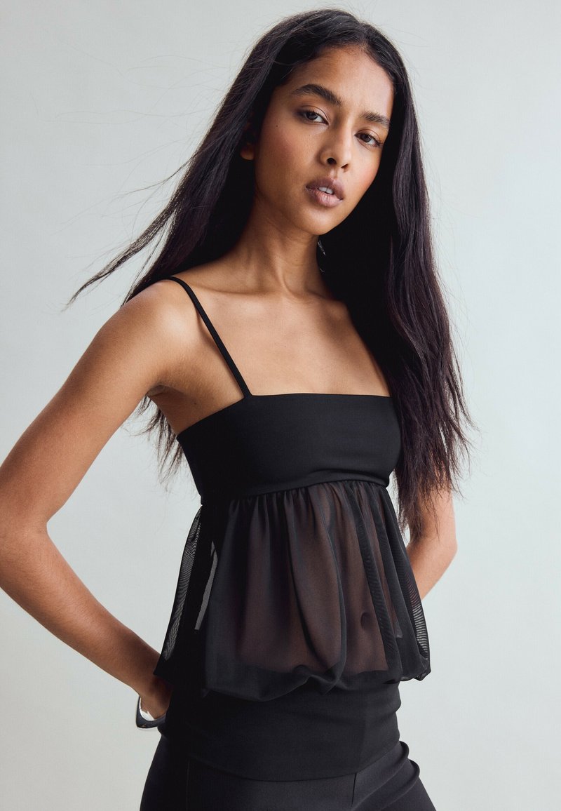 Femme aux longs cheveux bruns foncés portant un haut noir sans manches avec un voile en tissu transparent, debout, les mains sur les hanches, devant un fond uni.