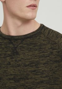 Mørk oliven grønn genser med en heathered tekstur. Har rund hals og kontrasterende svart søm langs kragen og skuldrene.