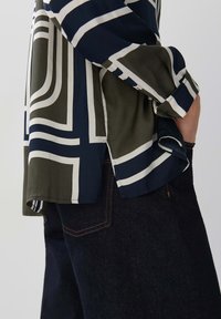 Het overhemd met lange mouwen heeft een marineblauw en olijfgroen ontwerp met witte geometrische strepen. Zacht materiaal met zijsplitten, gecombineerd met donkere spijkerbroeken.