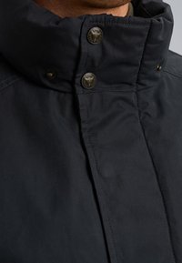 Mornarska vodoodporna jakna s povratnim zoomom, z visokim ovratnikom, ki ima dva medeninasta gumbka na pritisk in strukturirano tkanino.