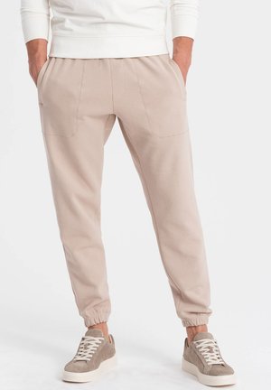 Pantaloni jogger beige realizzati in tessuto morbido con vestibilità rilassata, caratterizzati da due tasche frontali e polsini elastici, abbinati a sneaker grigio chiaro.