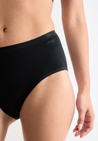 Torso femminile e parte alta della coscia in mutande alte nere con logo "Sloggi", su uno sfondo bianco uniforme.