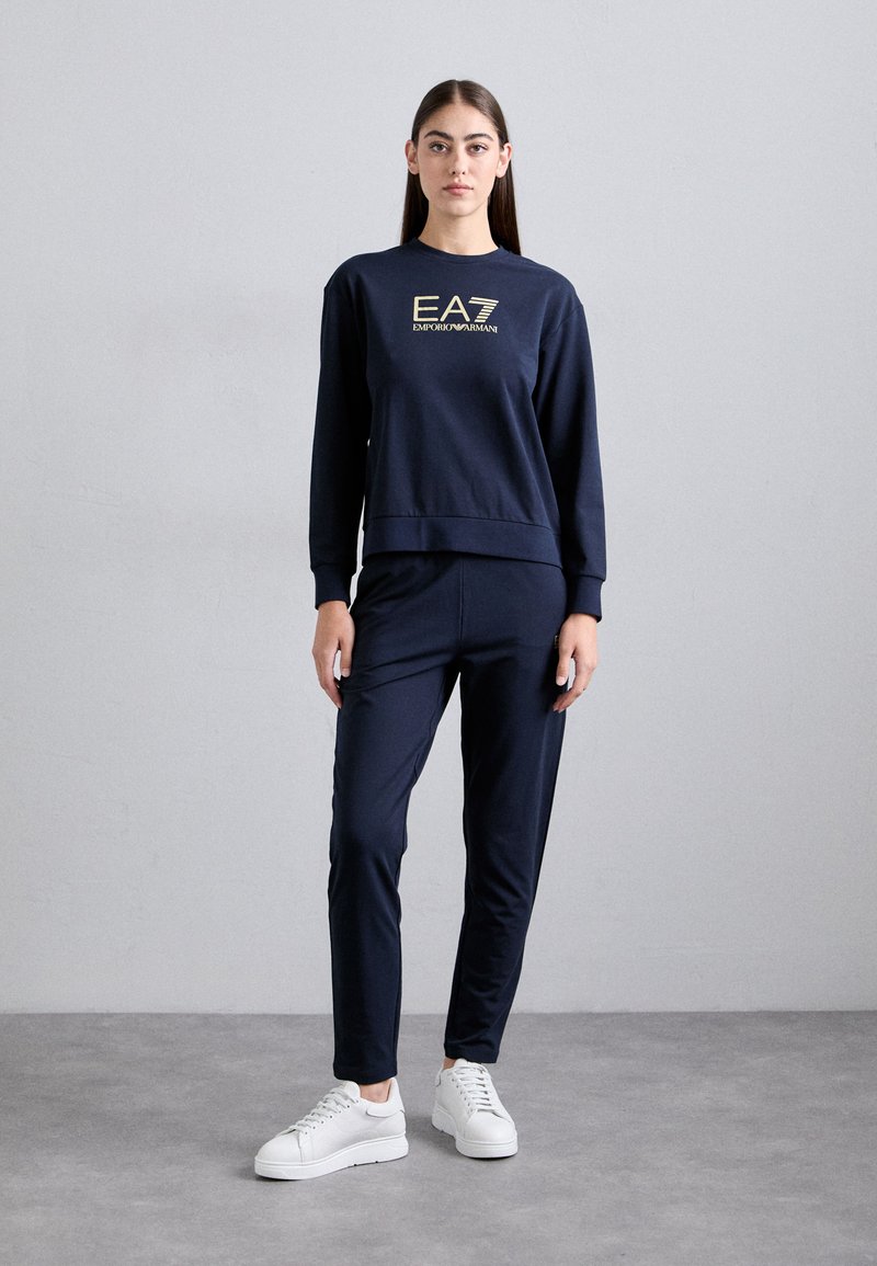 Tumši zili treniņi, kas sastāv no logotipa sporta krekla un pieguļošām biksēm, izgatavoti no mīksta auduma, kombinēti ar baltām sporta apaviem.