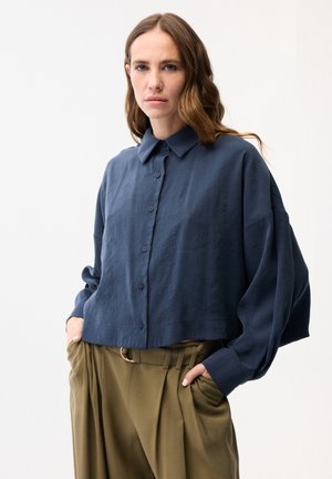 Kobieta o falujących brązowych włosach, ubrana w luźną, granatową koszulę zapinaną na guziki oraz oliwkowe, wysokowęższe spodnie z rękami w kieszeniach.