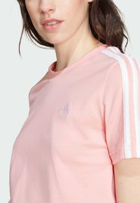 T-shirt de algodão cor-de-rosa com decote redondo, apresentando riscas brancas nos ombros e um pequeno logótipo bordado no peito.