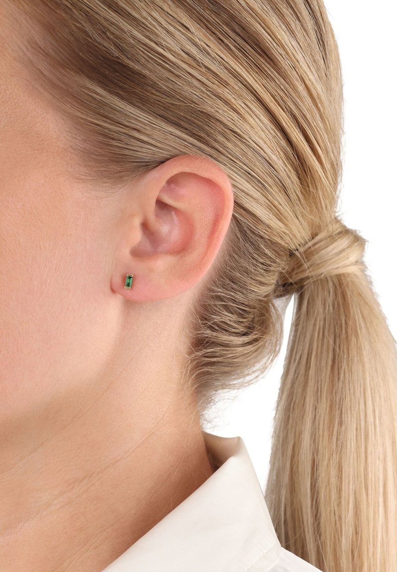 Donna con capelli biondi raccolti, che indossa un piccolo orecchino a goccia di pietra verde rettangolare nell'orecchio sinistro, su uno sfondo bianco.