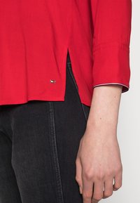 Maglietta rossa leggera a manica lunga con spacco laterale, caratterizzata da un piccolo logo e cuciture a contrasto sul polsino.