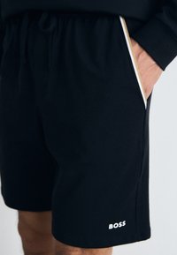 Svart shorts laget av myk stoff med snor i livet, side lommer med hvite og gull detaljer, og en hvit "BOSS" logo.