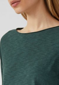 T-shirt vert foncé, texturé, avec un large col rond. Tissu doux avec un motif chiné s'étendant sur le matériau.